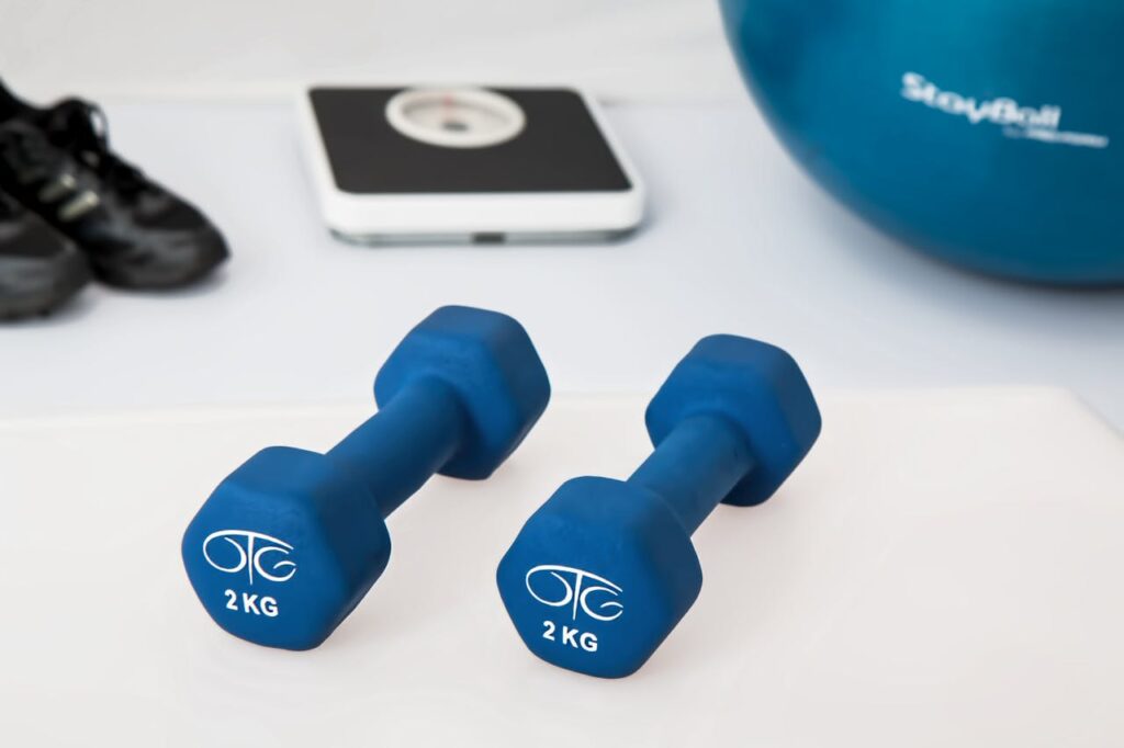 physiotherapy-weight-training-dumbbell-exercise-balls-39671 Blue dumbbells placed on a white surface with fitness equipment in the background.